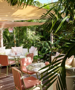 Two outdoor dining tables surrounded by greenery with pink chairs and a tan shade hanging above