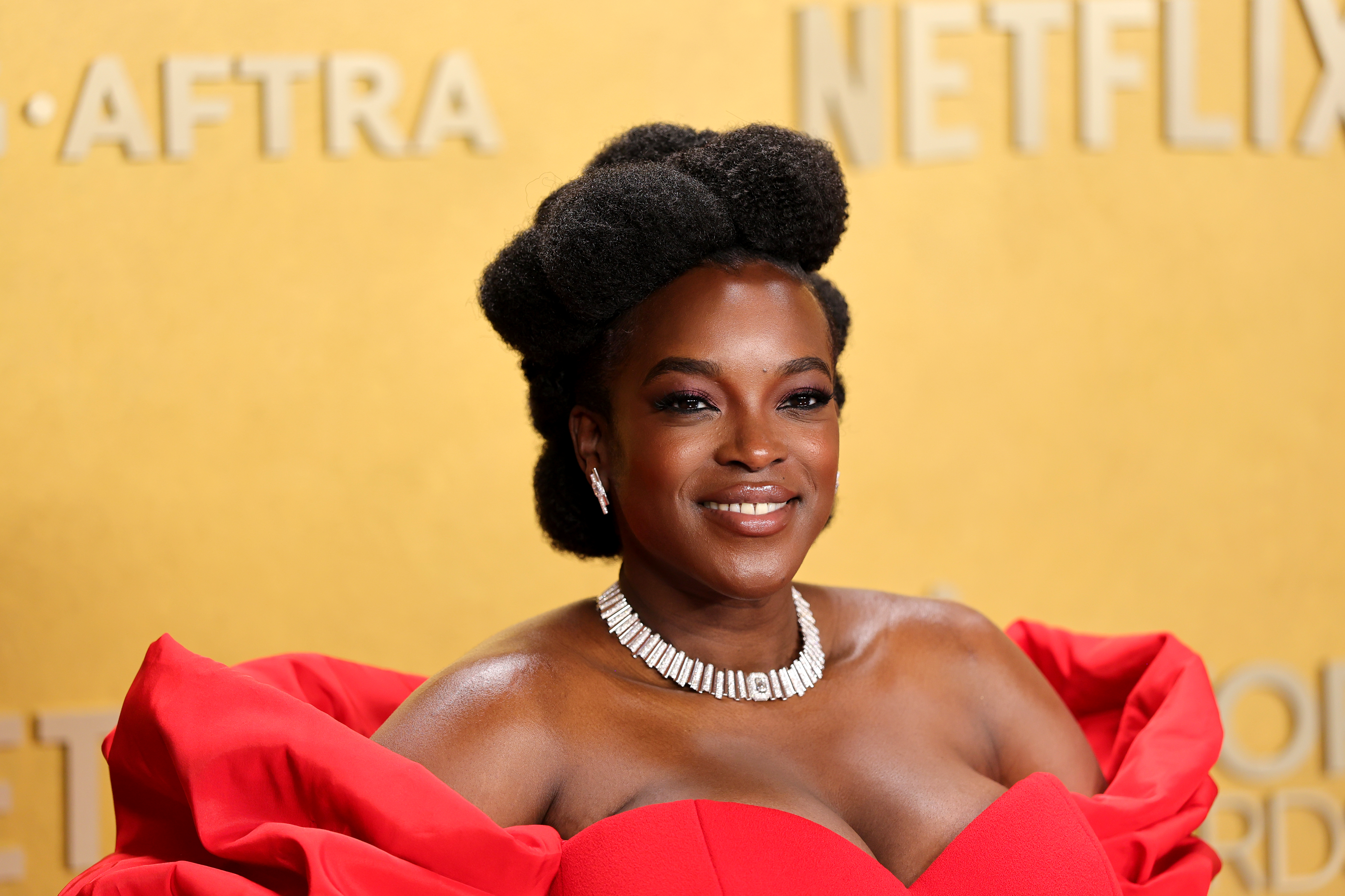 a black woman with textured hair wears an updo and a red gown while posing in front of a yellow backdrop
