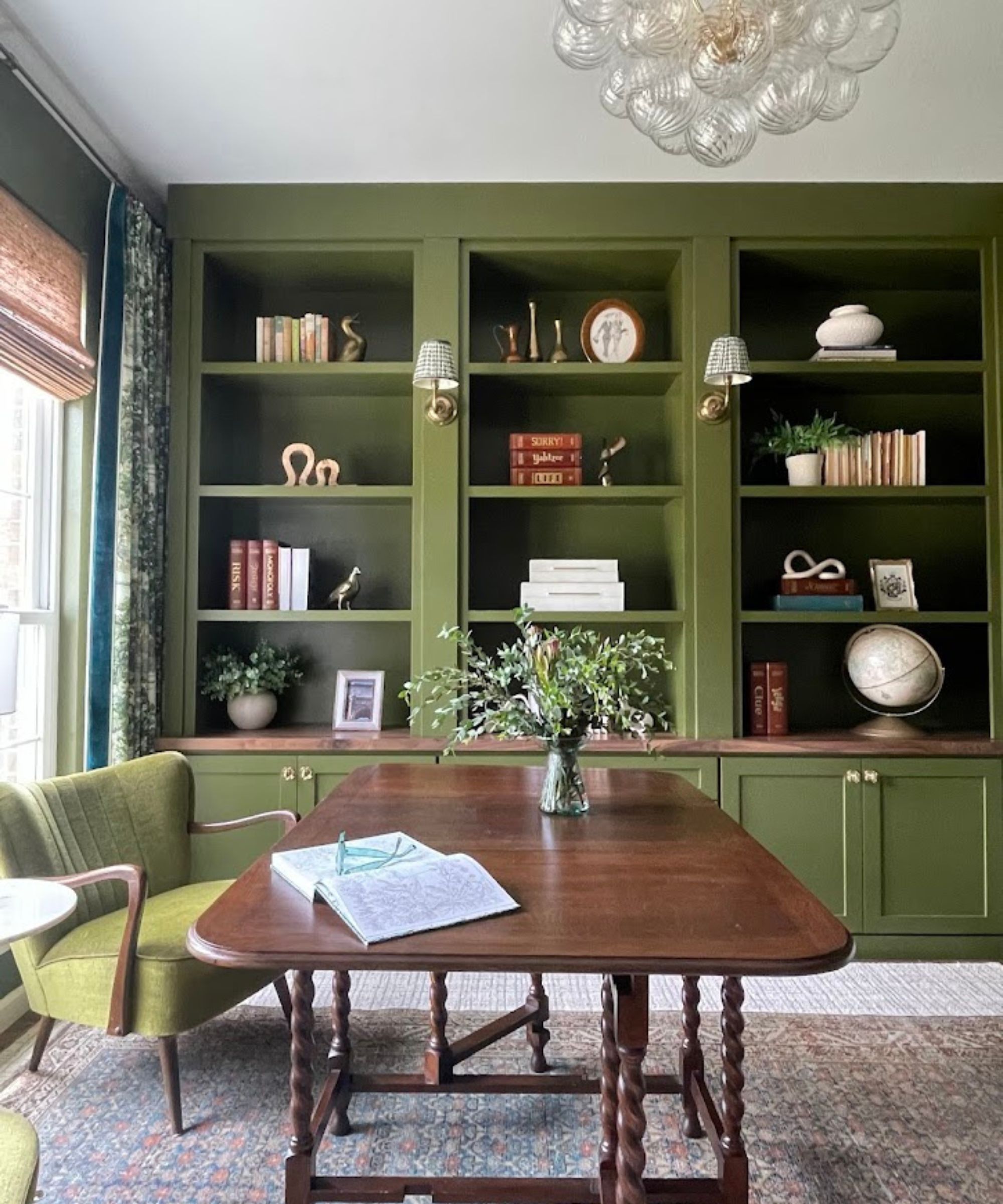 Library with dark wood table, green armchair, green painted shelving unit and sconces and large rug