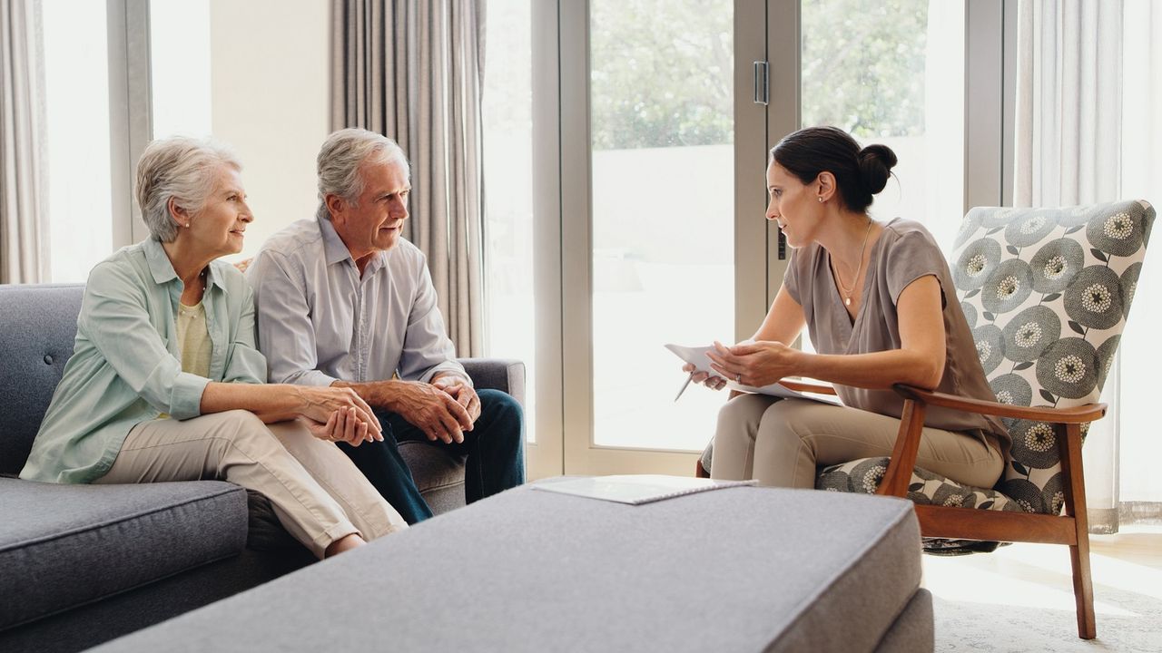 A retired couple sit on a couch talking with a financial adviser.