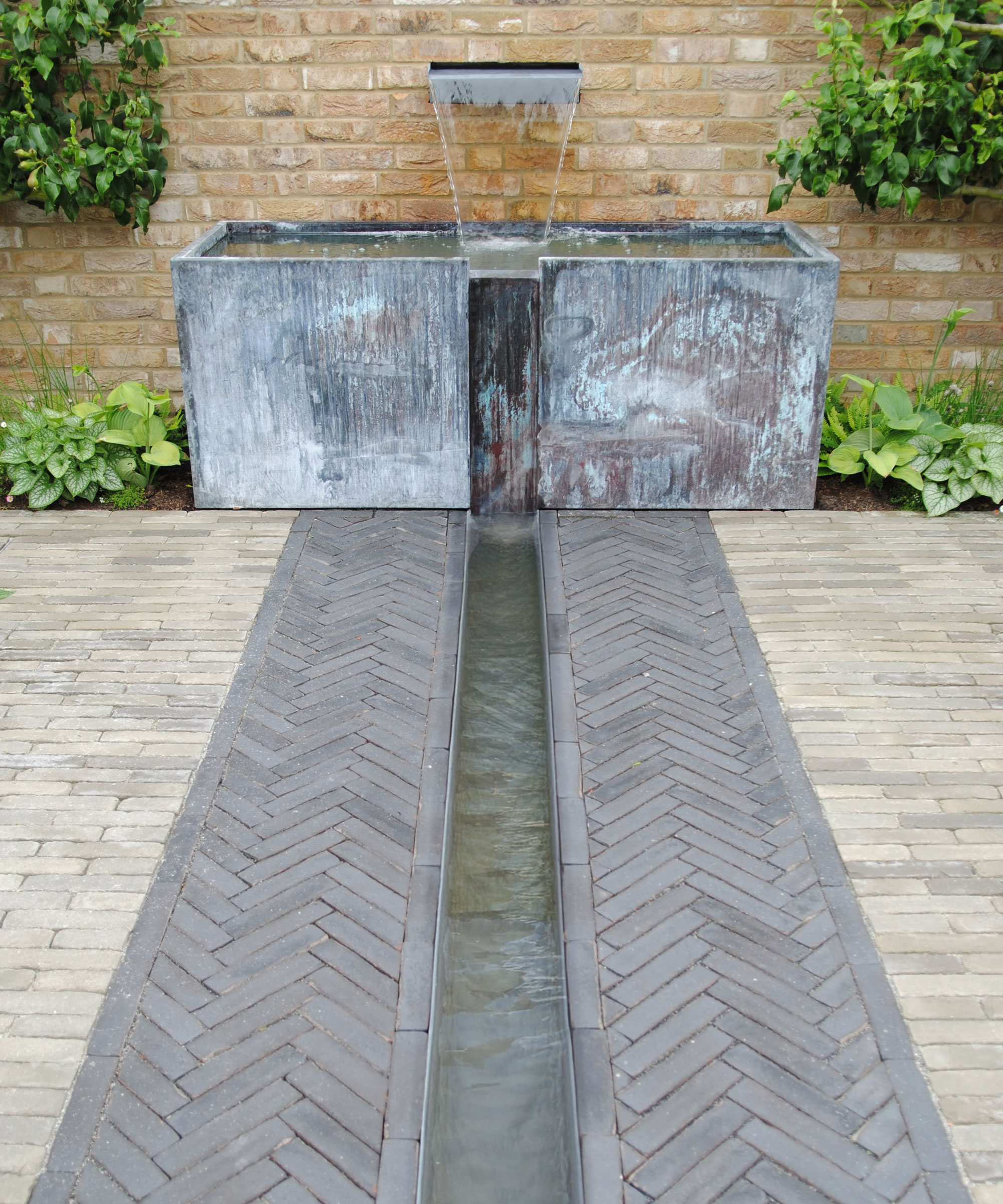 galvanised water feature with water blade and tank, set against a brick wall, with a rill and herringbone pattern paving and plantin