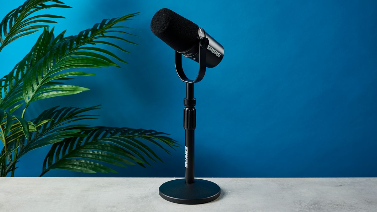 a black microphone with a desktop stand and a black muff is on top of a beige surface with a blue backdrop