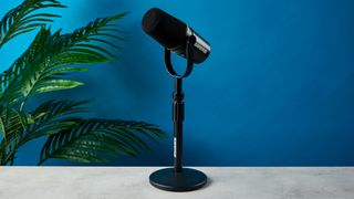 a black microphone with a desktop stand and a black muff is on top of a beige surface with a blue backdrop