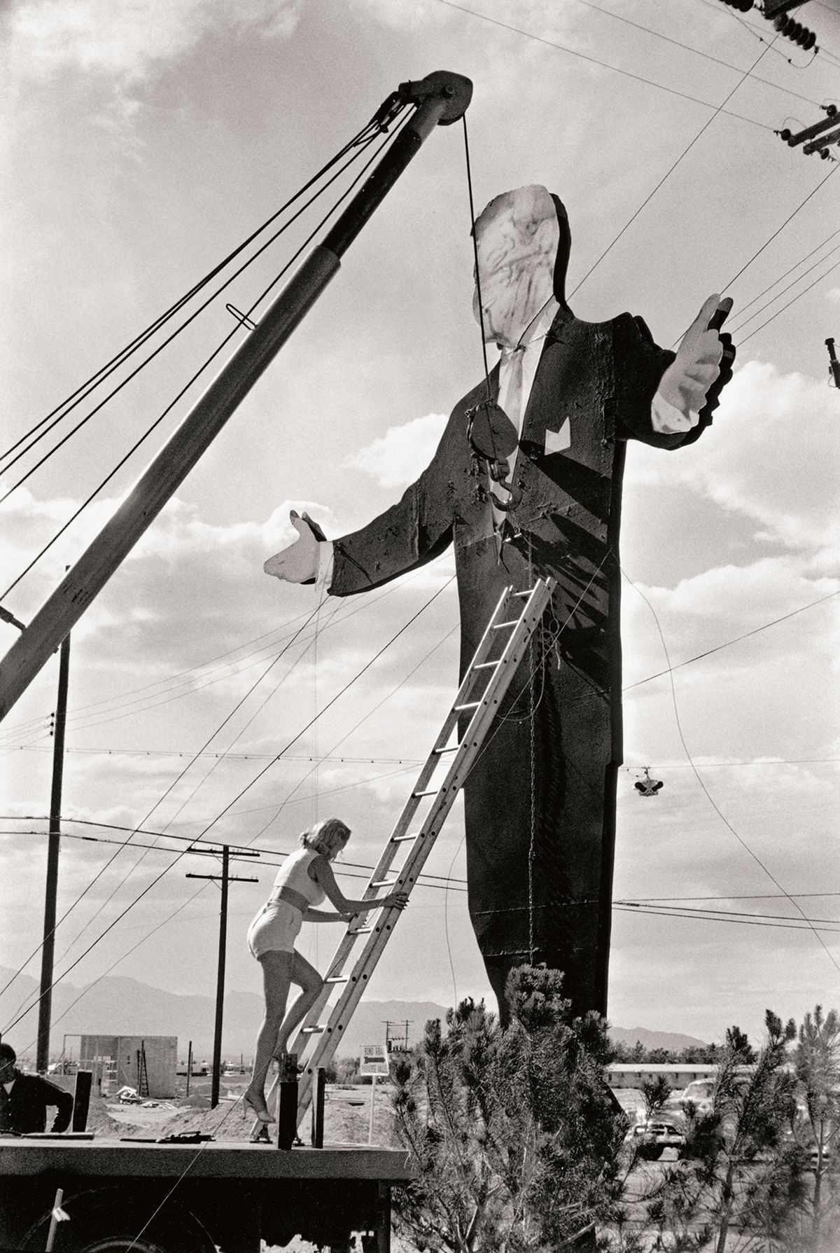 A black and white image of a woman on a ladder works on a giant statue of a man in a suit, under a cloudy sky with power lines in the background
