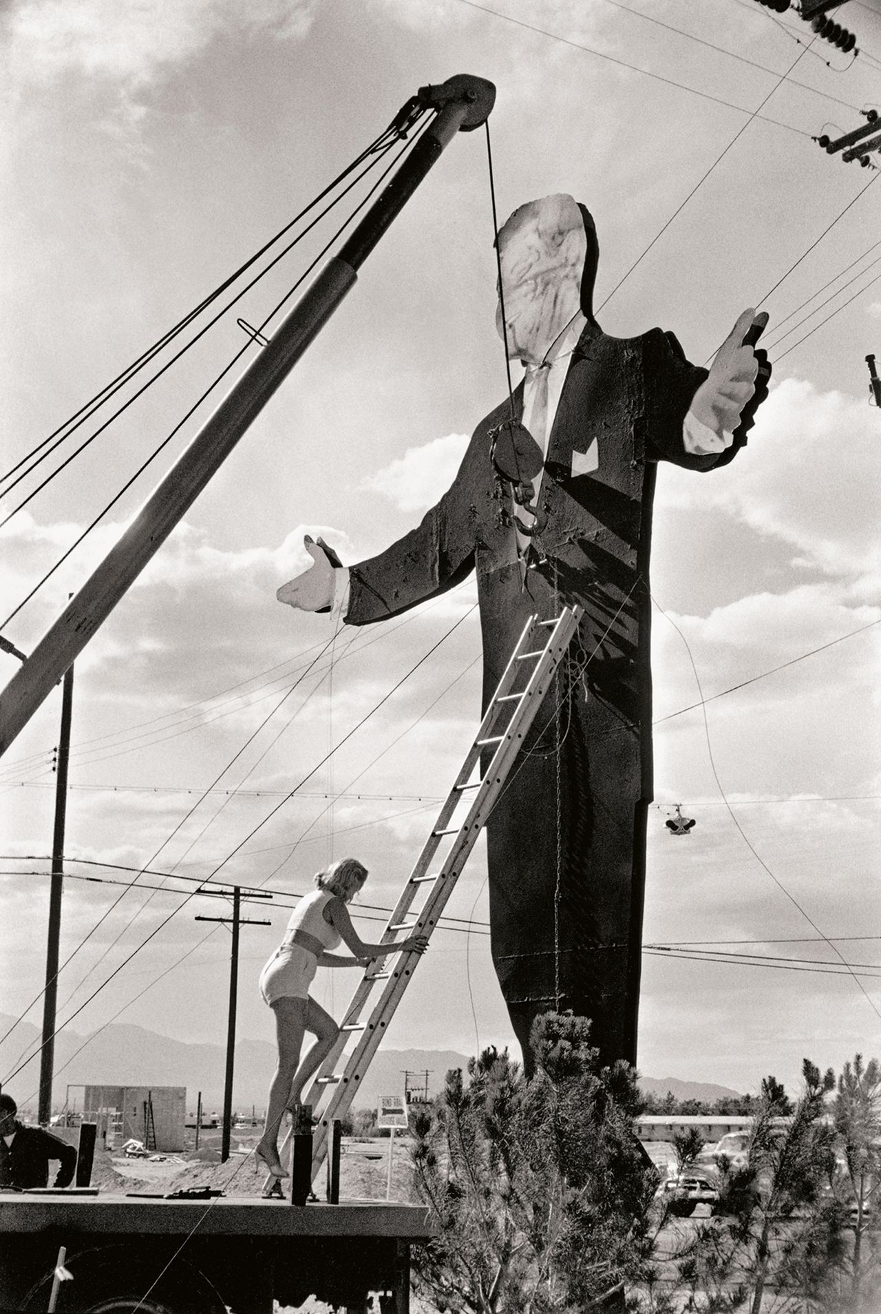 A black and white image of a woman on a ladder works on a giant statue of a man in a suit, under a cloudy sky with power lines in the background