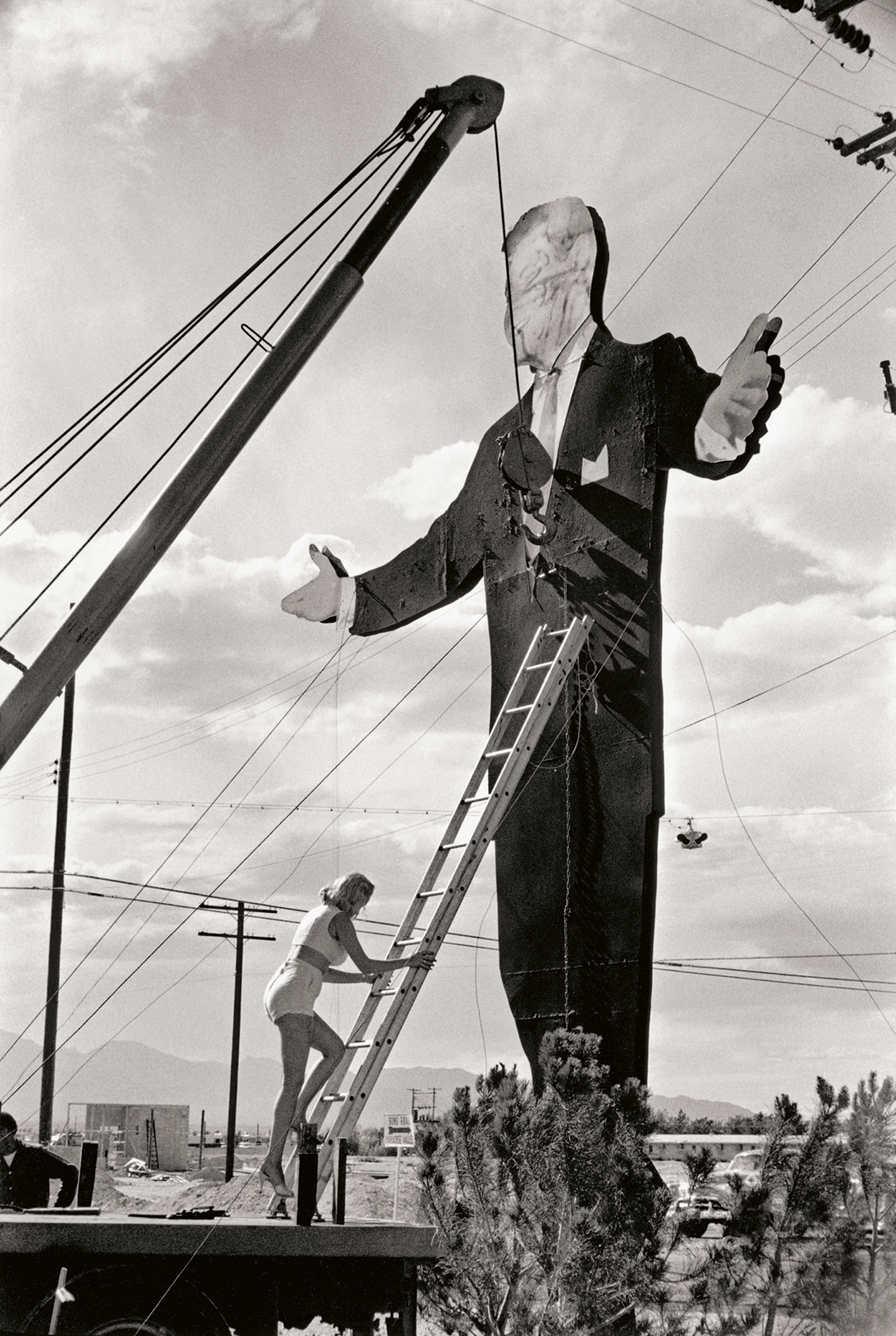 A black and white image of a woman on a ladder works on a giant statue of a man in a suit, under a cloudy sky with power lines in the background
