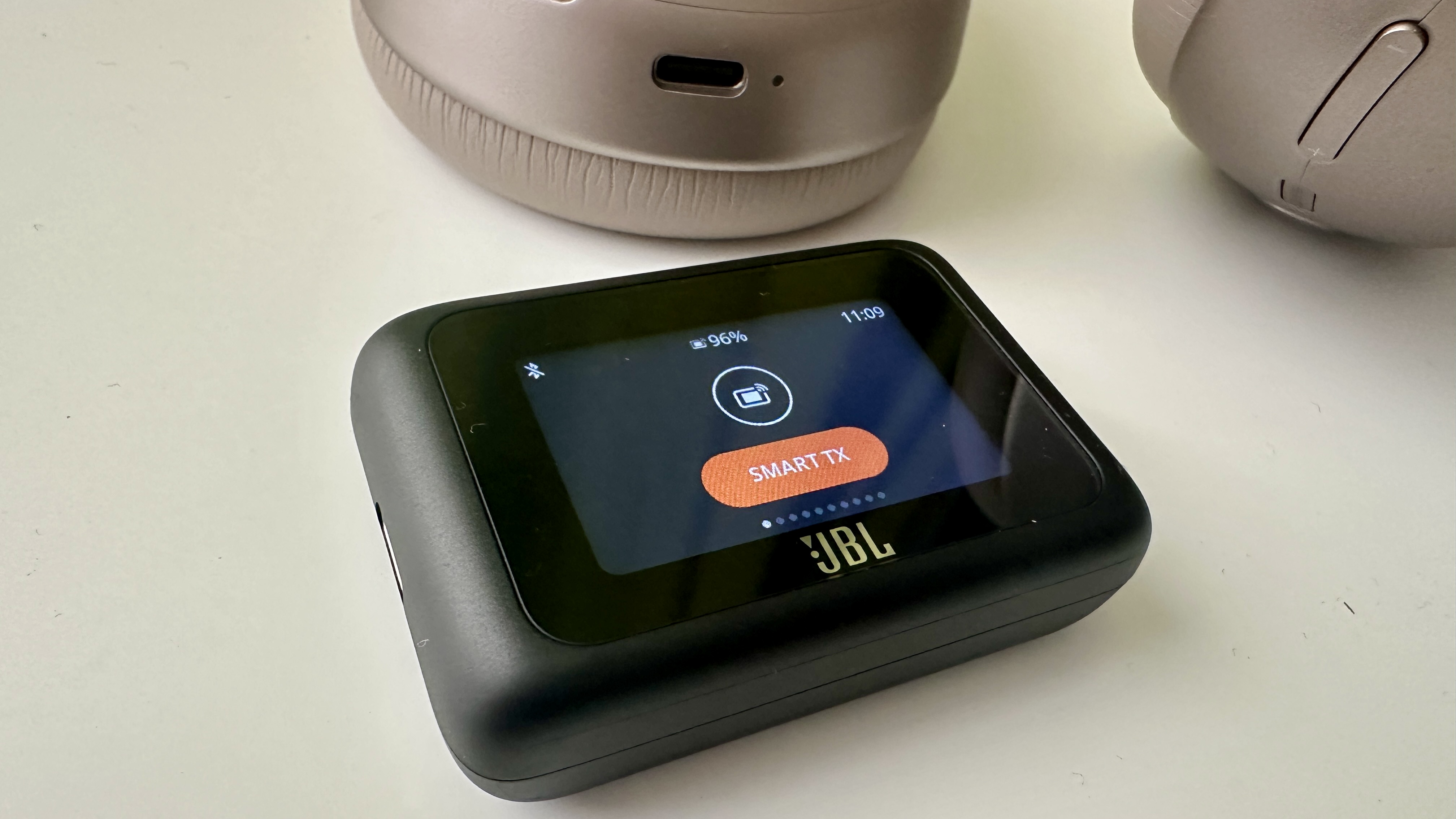 Optional ‘Smart Tx’ transmitter for the JBL Tour One M3 headphones, both on a white surface
