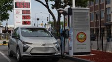 An electric Chevy vehicle charges in front of a gas station with high prices. 