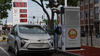 An electric Chevy vehicle charges in front of a gas station with high prices. 