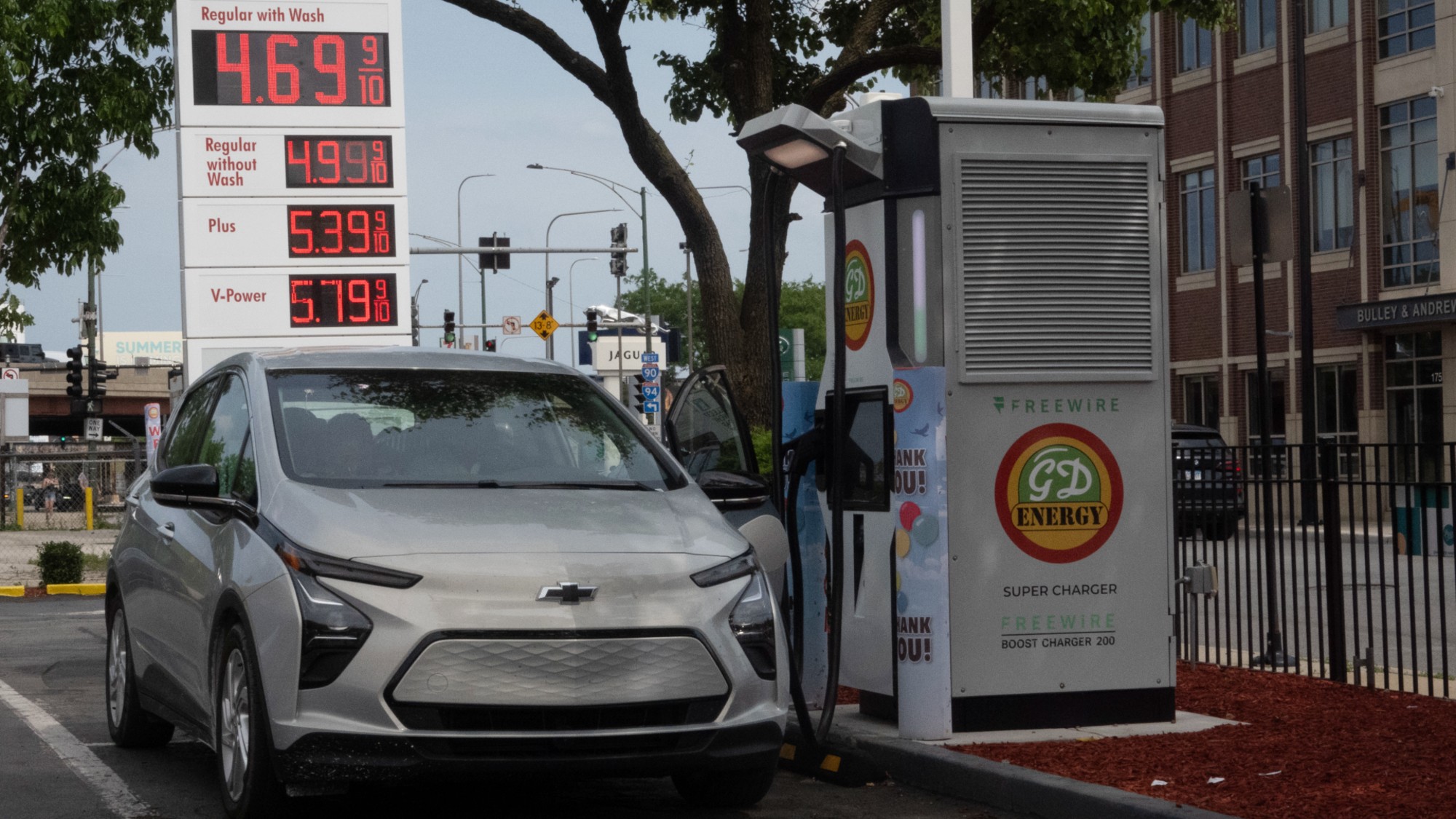 An electric Chevy vehicle charges in front of a gas station with high prices.  