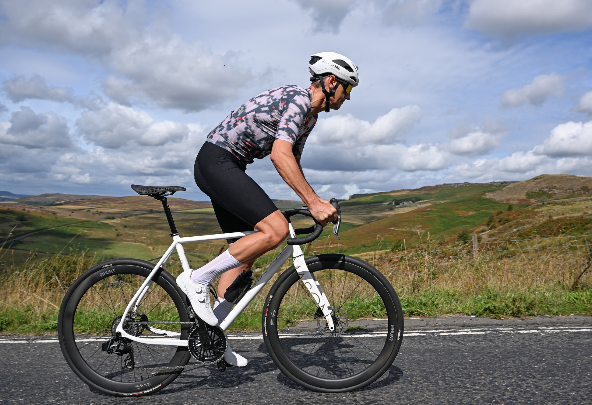 a white road bike being ridden out of the saddle by a man wearing white shoes, white helmet, black shorts and a patterned grey jersey with countryside hills in the background