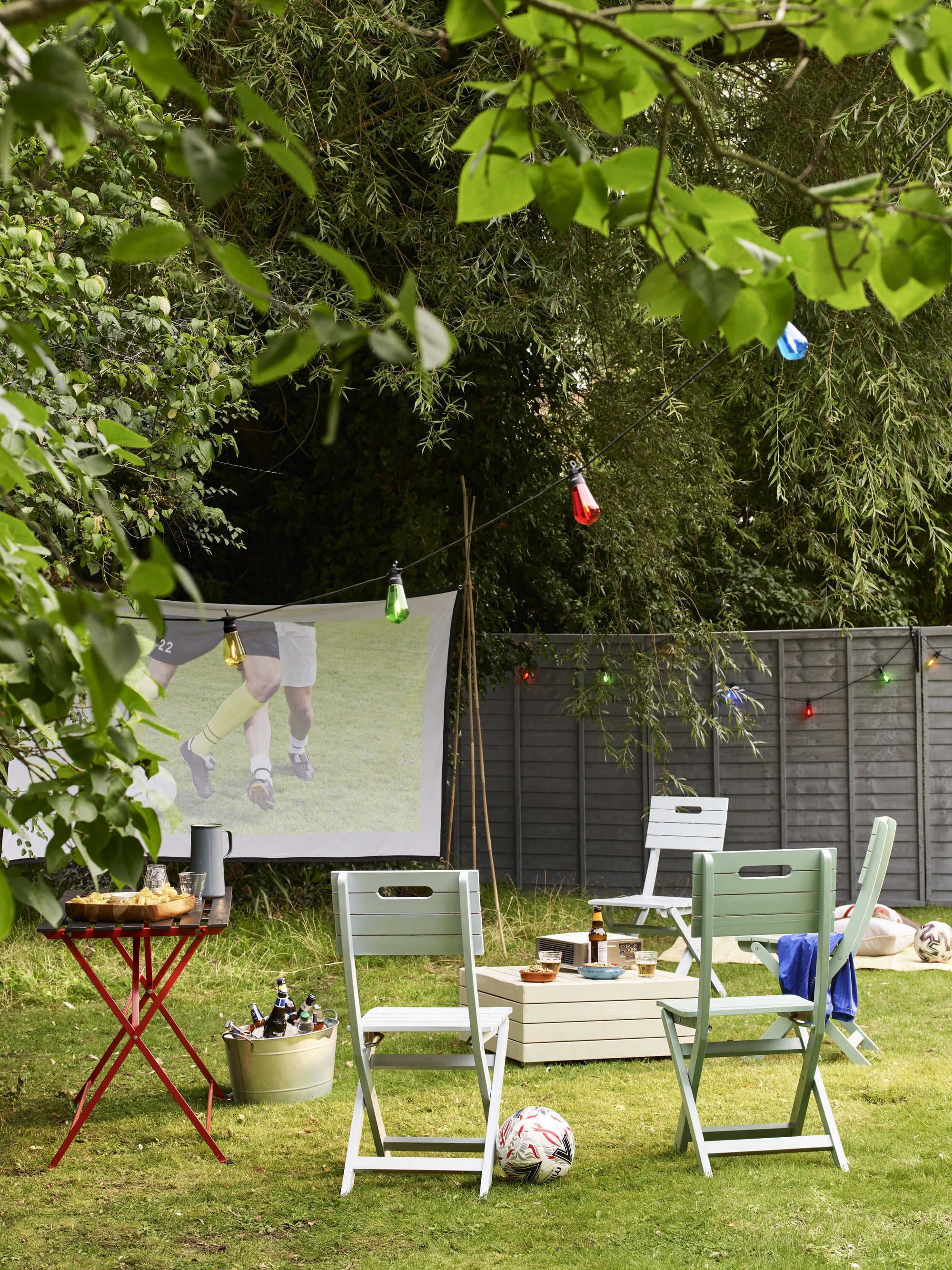 Image of a grassy backyard with lots of tree coverage and a projector screen hanging in the back corner by a gray fence. There are wooden folding chairs painted teal positioned around a wooden coffee table and in front of the screen.