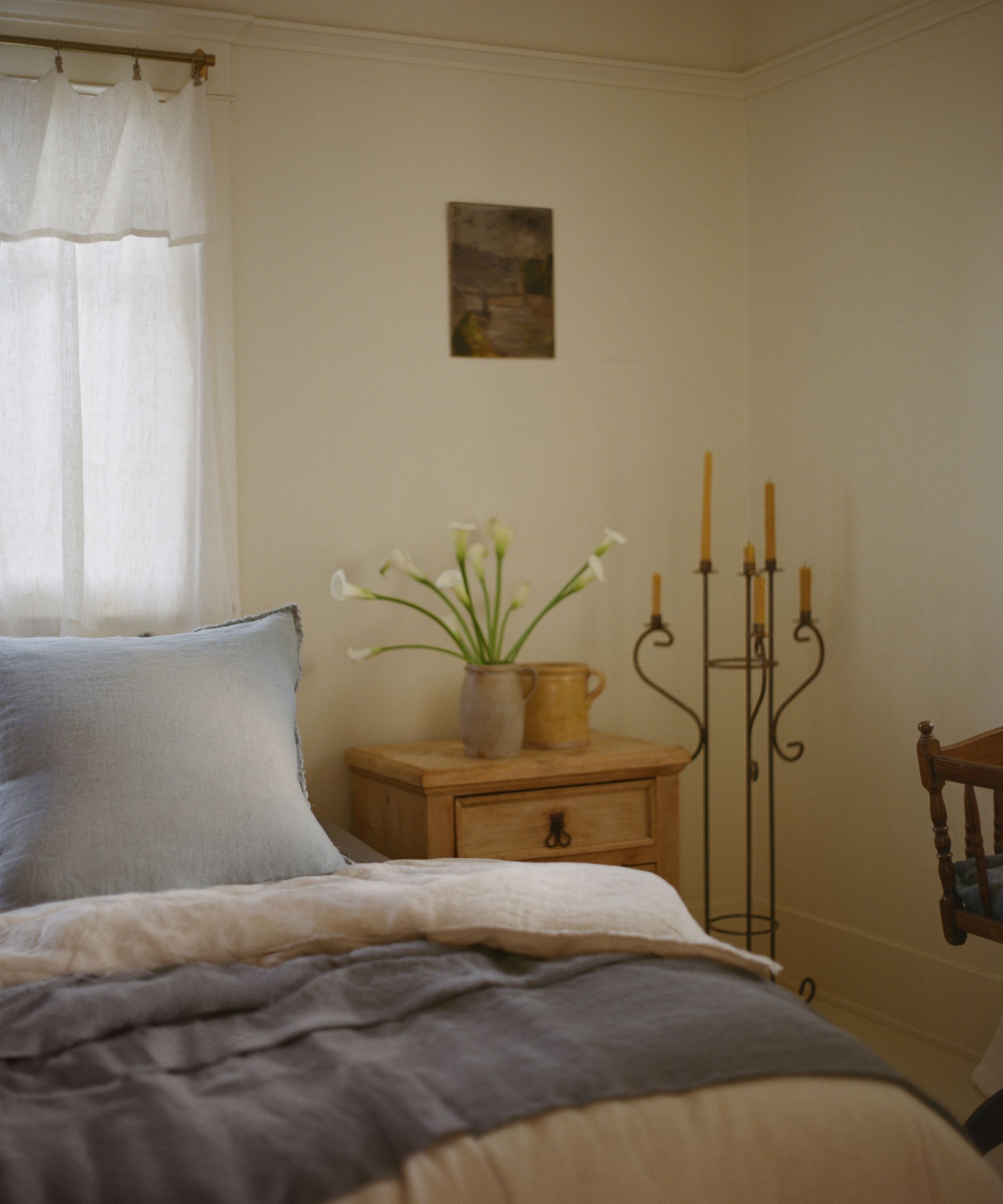 Cozy bedroom corner with a bed in soft grey linen, a light wood nightstand with calla lilies, and a tall iron candelabra.