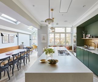 large kitchen diner with large cream island in middle of room, dark green units and coper splashback on one side and long refectory style dining table on other with rooflights above