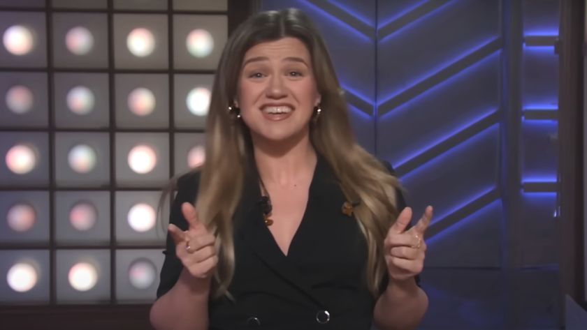 Kelly Clarkson stands smiling with finger guns in front of her set on The Kelly Clarkson Show.