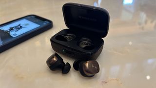 The Sennheiser Momentum True Wireless 4 headphones out of their case, sat next to a phone