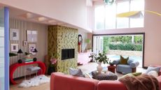 A pale pink living room with a colorful tiled wall, a reddish pink curved couch, blue plush sofa seats, a patterned stacked coffee table seat, a red curved side table with books, plants, matching lamps, a metal stool and a furry rug in front of a striped accent wall