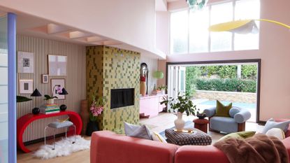 A pale pink living room with a colorful tiled wall, a reddish pink curved couch, blue plush sofa seats, a patterned stacked coffee table seat, a red curved side table with books, plants, matching lamps, a metal stool and a furry rug in front of a striped accent wall