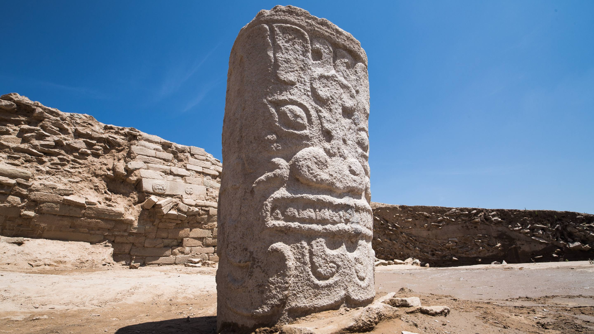 a large rock carved with a face stands in front of archaeological ruins of a wall