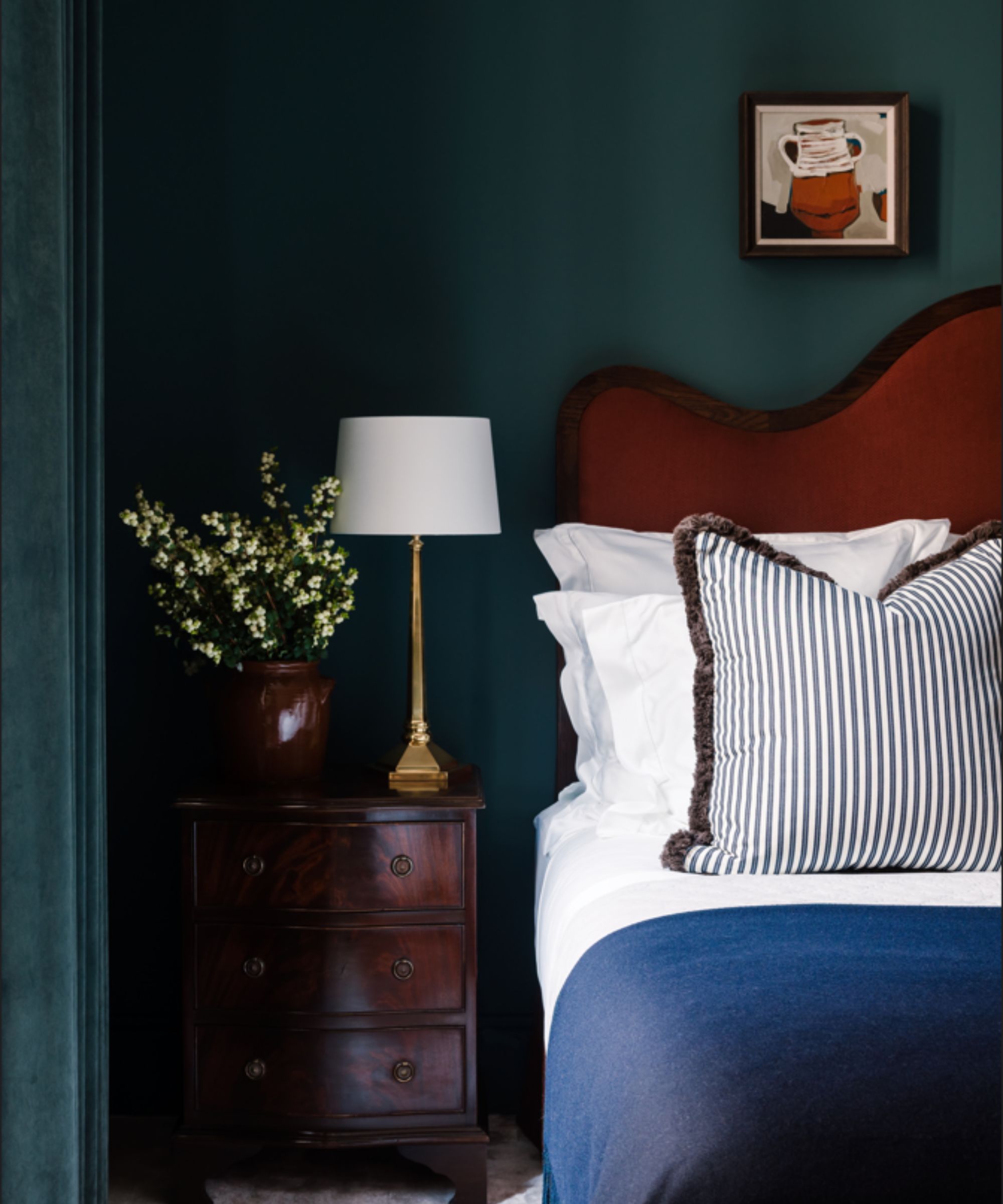 A deep blue bedroom with a red velvet bed, and a dark wood oak side table.