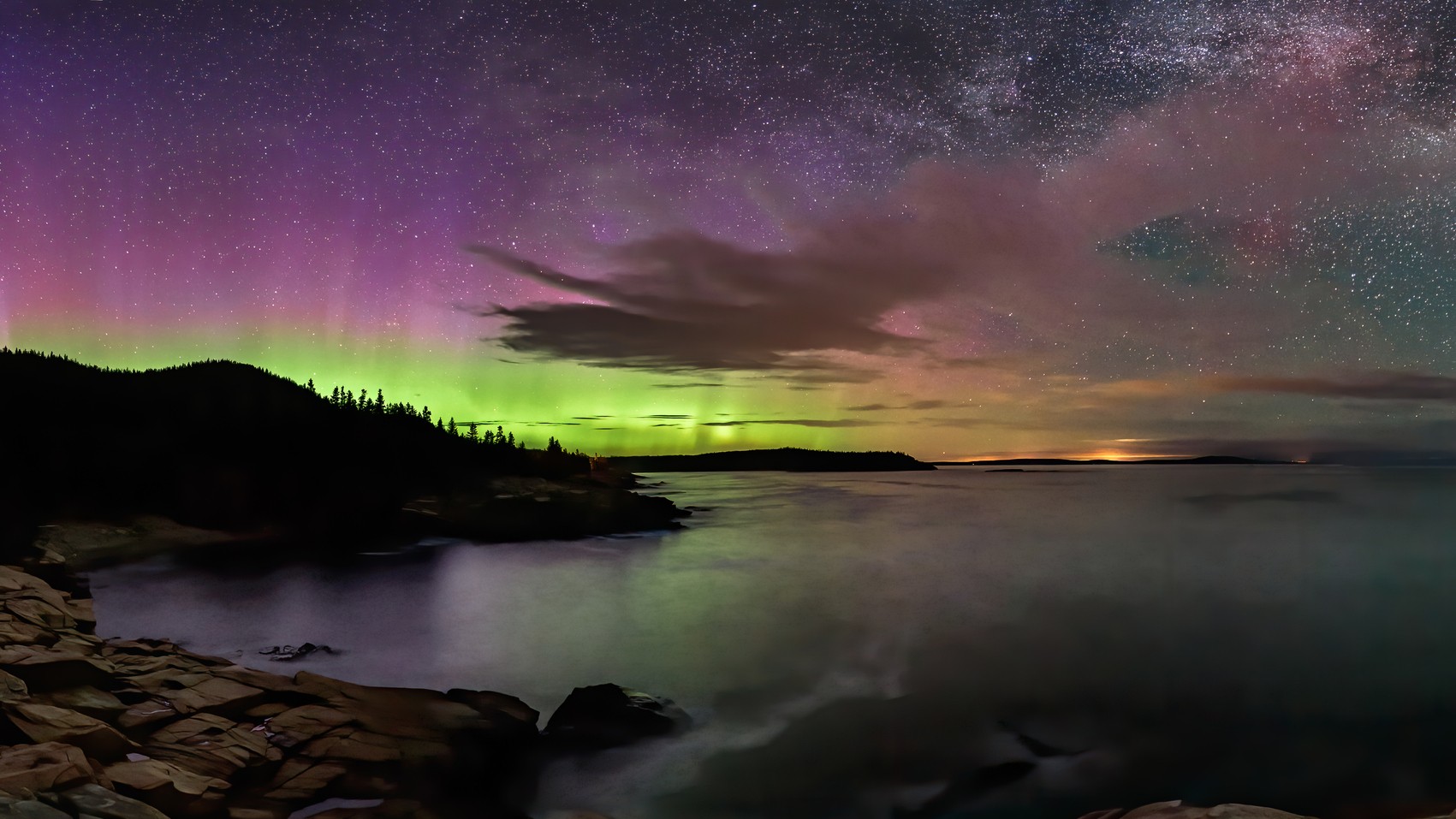 northern lights and starry sky shine above water below and trees silhouetted in the distance. the northern lights appear as curtains of green and purple light. 