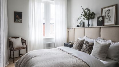 bedroom curtain mistakes, neutral bedroom with padded oatmeal headboard, taupe bedding, patterned cushions, shelf above headboard, vintage chair in corner, white voile curtains, radiator, wooden floor boards, artwork 