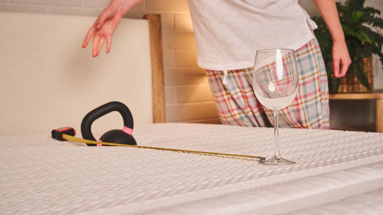 A person drops a kettlebell onto the surface of the Leesa Legend Hybrid Mattress. An empty wine glass sits upright 25 inches away from the weight, with a tape measure showing the distance between the two