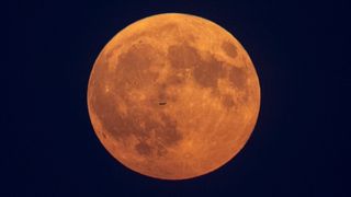 The silhouette of a plane flies in front of a full moon with a very orange hue