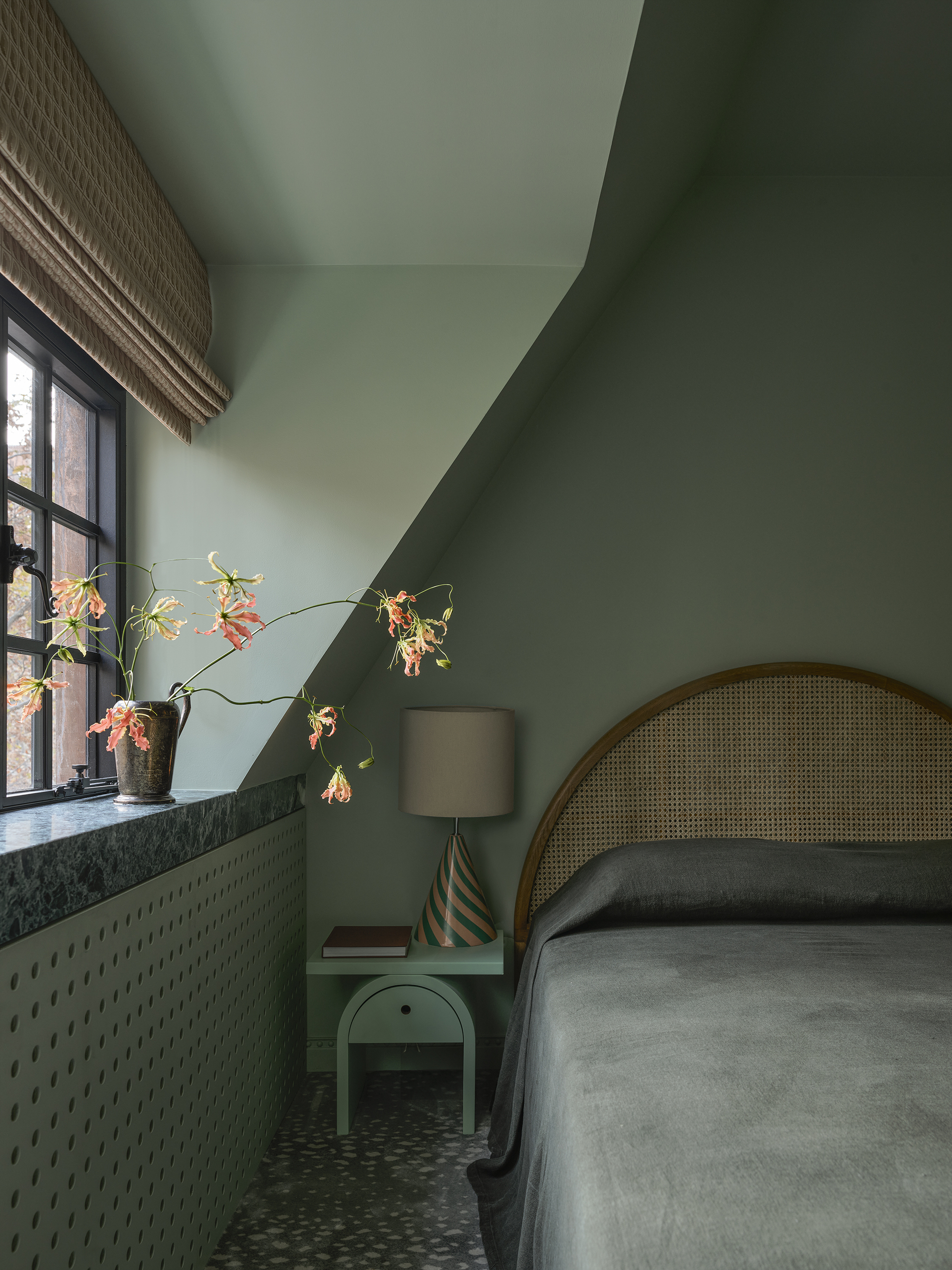 green color-drenched bedroom with green marble windowsill, roman blinds, rattan bedhead, green side table with striped lamp, and green bedding