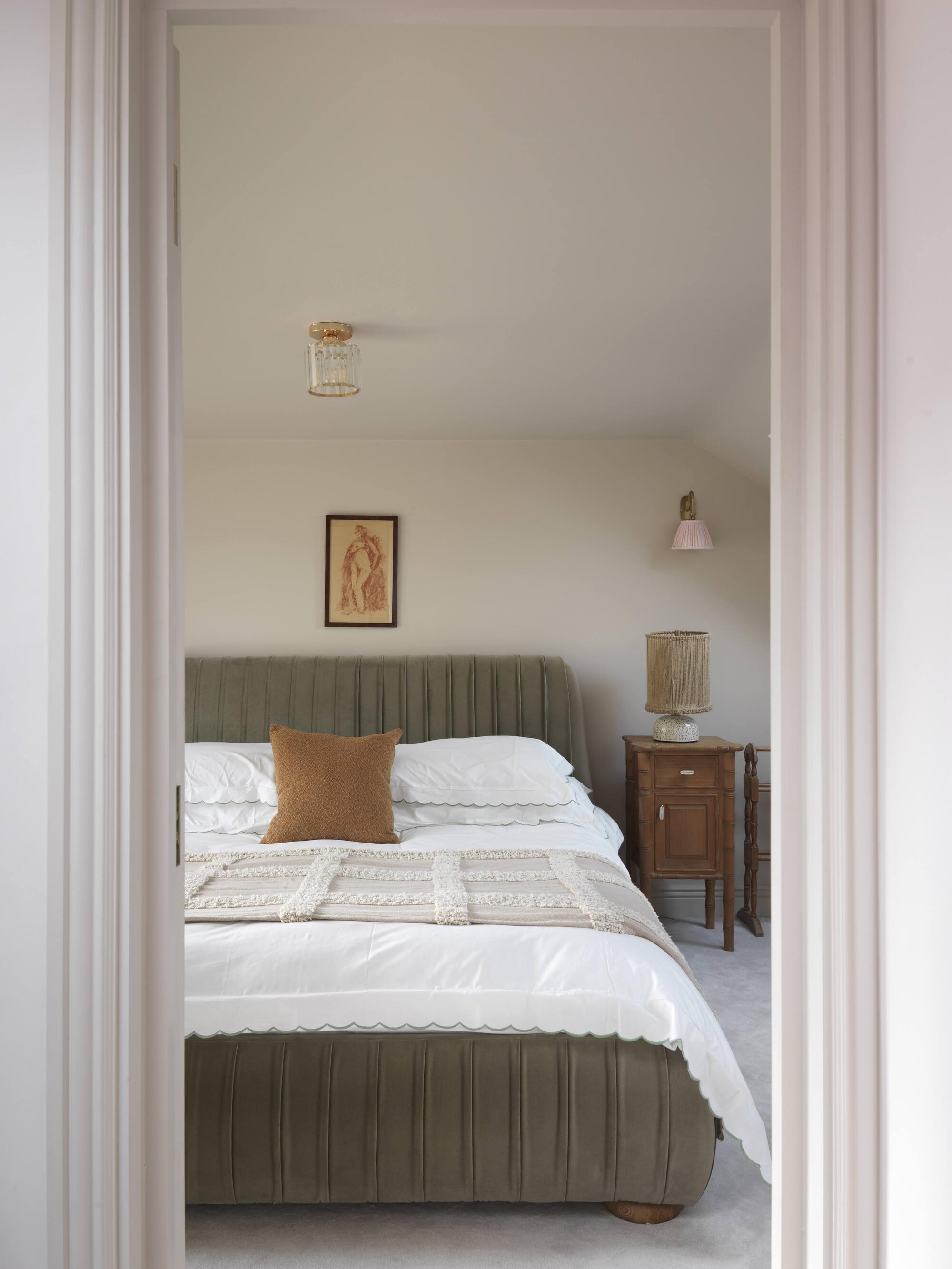 earth toned bedroom with blush walls, green bed, white sheets, timber side table, artwork over bed, ceiling light, and wall sconce