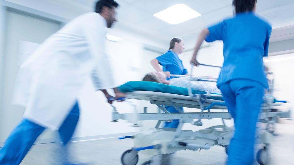 three doctors in white coats and scrubs quickly pushing a patient on a bed down a hall of an emergency department