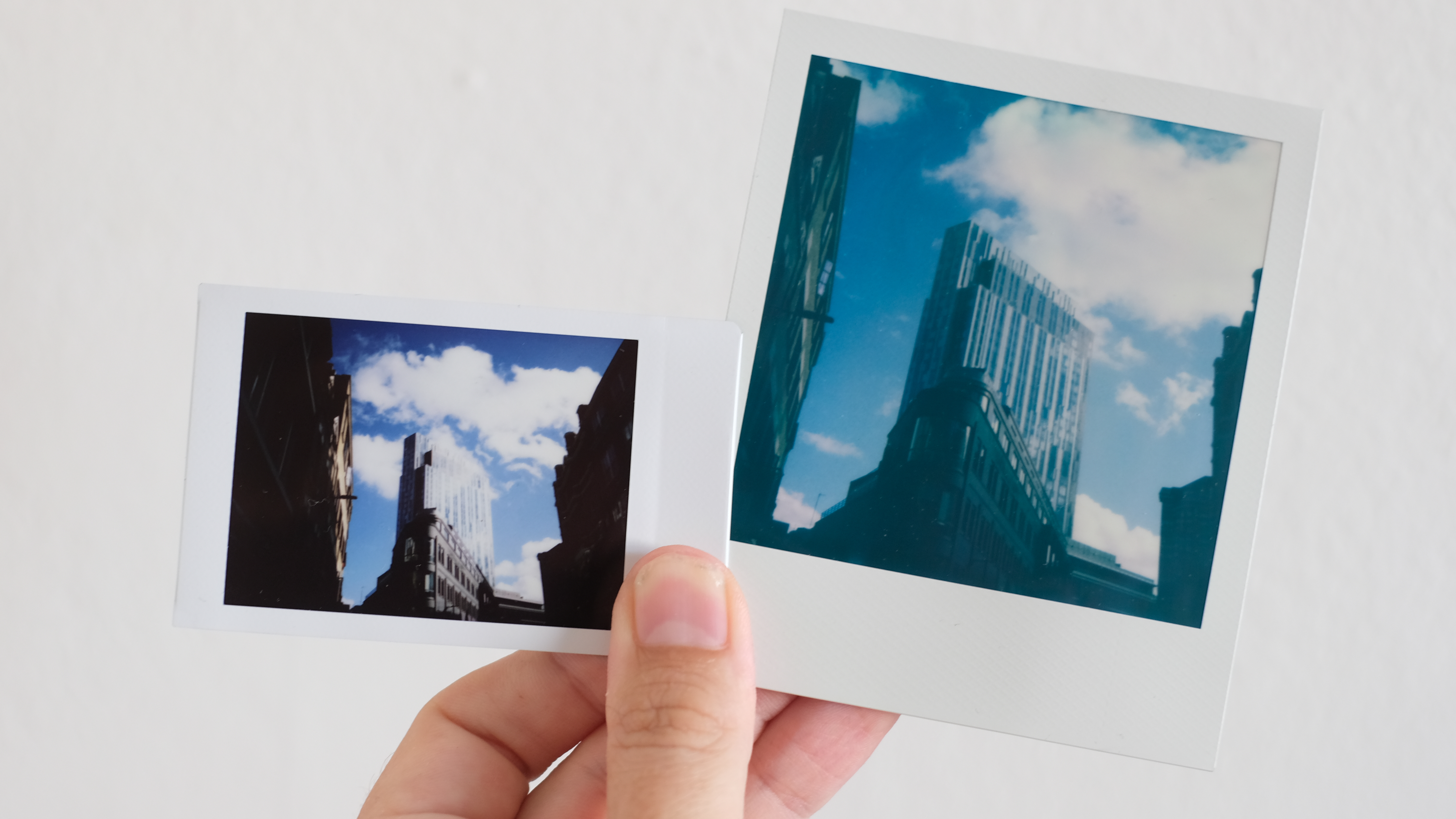 Mano sosteniendo imágenes de Instax y Polaroid mostrando el mismo edificio; Instax es más brillante y dramático, pero Polaroid tiene mejor detalle y gradaciones tonales sutiles