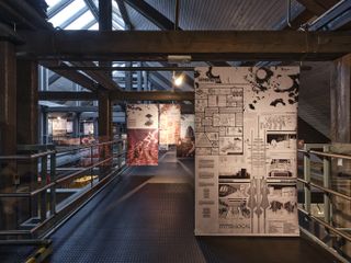 Interior view of the exhibition panels at tallinn architecture biennale 2022, dark wood ceiling beams, arched glass skylight windows, silver handrail, black flooring, lighting