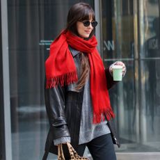 Dakota Johnson walks outside wearing black straight-leg jeans with loafers, a grey knit, a black leather jacket, a red scarf and a leopard print bag.