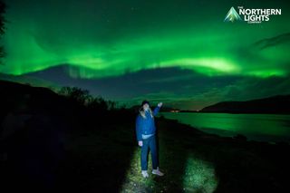 a woman stands in front of a green sky filled with auroras and holds out her hand to point "there they are"