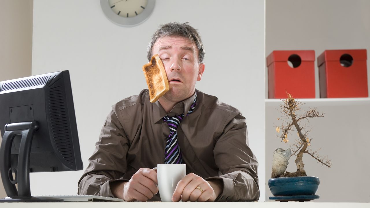 A man sitting at his desk looks like he just woke up, a piece of toast sticking to his face.