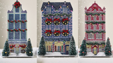 three anthropologie christmas houses in blue purple and pink with glitter and fake snow falling in the background with two small frosted christmas trees either side of the door of each house