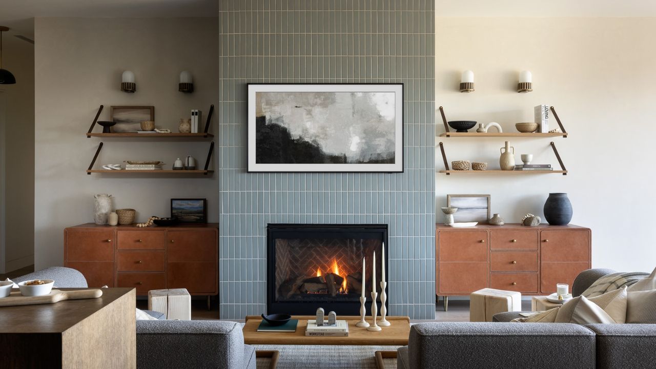 Living room with blue tiles and white walls, with floating shelves with various ornaments, a framed painting, a fireplace, two gray sofas, and a coffee table with books and candlesticks.