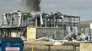 the skeleton of a building stands smoking in rubble after a fire. A blue dumpster in the foreground on the bottom left has a sign that reads 'NO PARKING'
