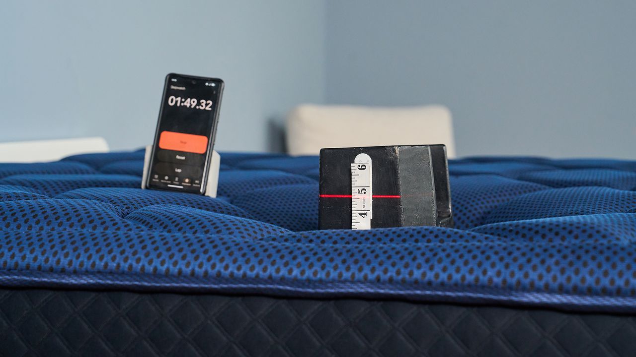 The image shows a 25kg weight placed on the surface of a dark blue mattress, with a red laser level tracking its level of sink. In the background, a phone screen displays a stopwatch.