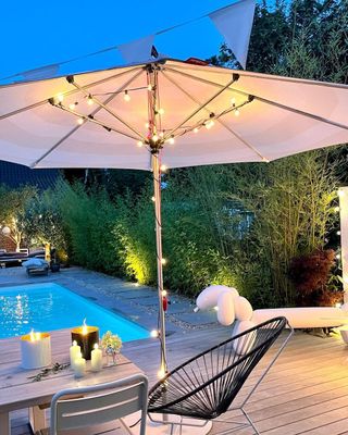 Umbrella with string lighting, chairs, decking and a pool in the background