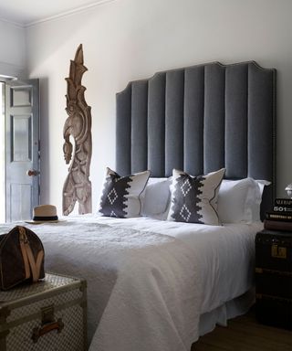 Bedroom with tall, gray upholstered headboard, white bedding, black and white geometric cushions, wooden sculpture next to bed, dark wood flooring, storage chest at end of bed
