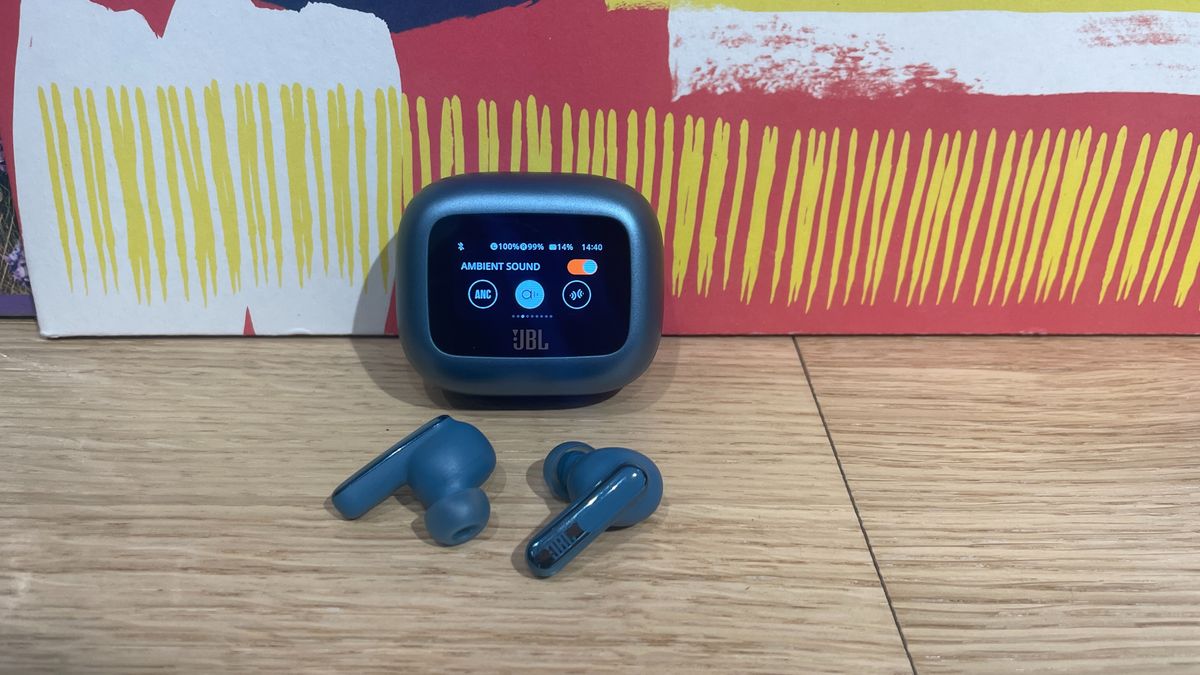 The blue case for a pair of JBL Live Beam 3 earbuds. The lid is closed and it is propped up with the screen on the front visible. Both of the buds have been removed and placed on the wooden surface in front of the case.