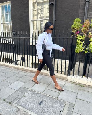 Marilyn Nwawulor-Kazemaks wearing a white button-down shirt with capri pants, strappy kitten heel sandals, and a black Alaïa Le Teckel bag.