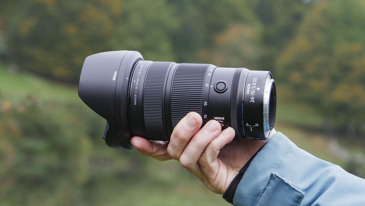Nikon Z 24-70mm f/2.8 S II lens in user's hand, with autumnal lake background