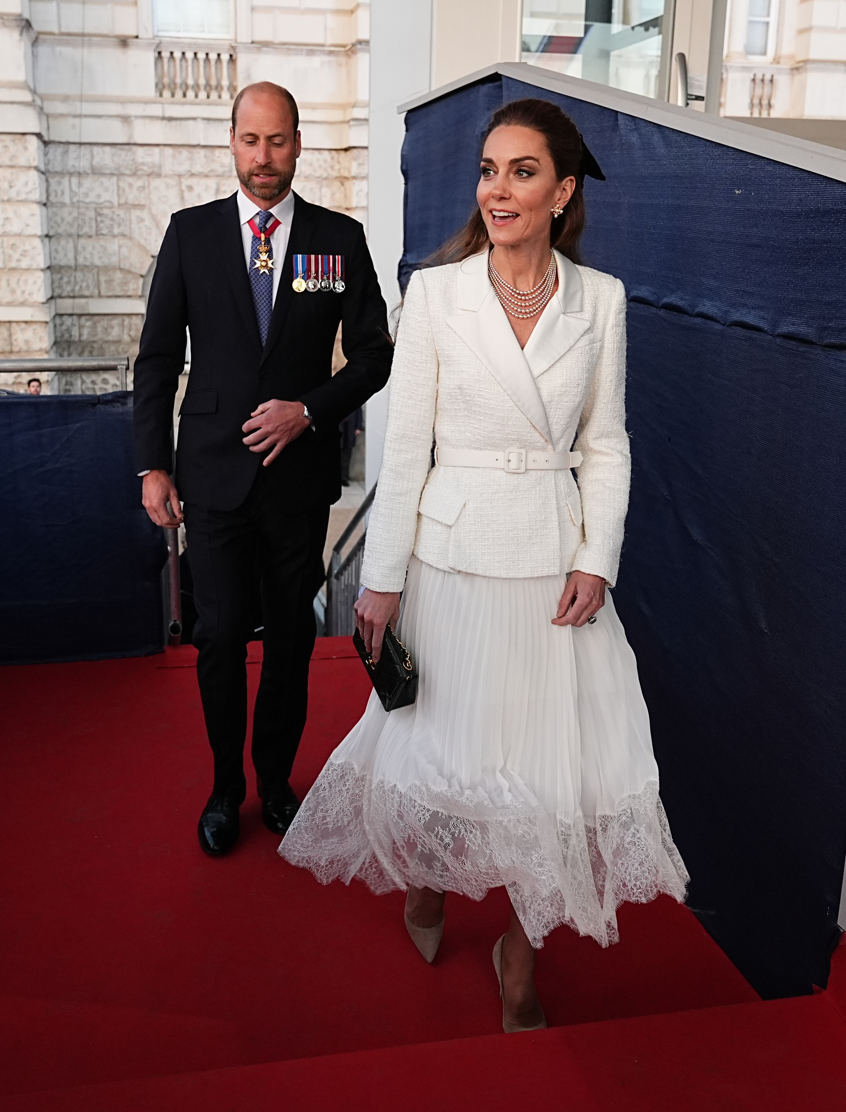 Kate Middleton wearing a white blazer dress walking down a red carpet with Prince William