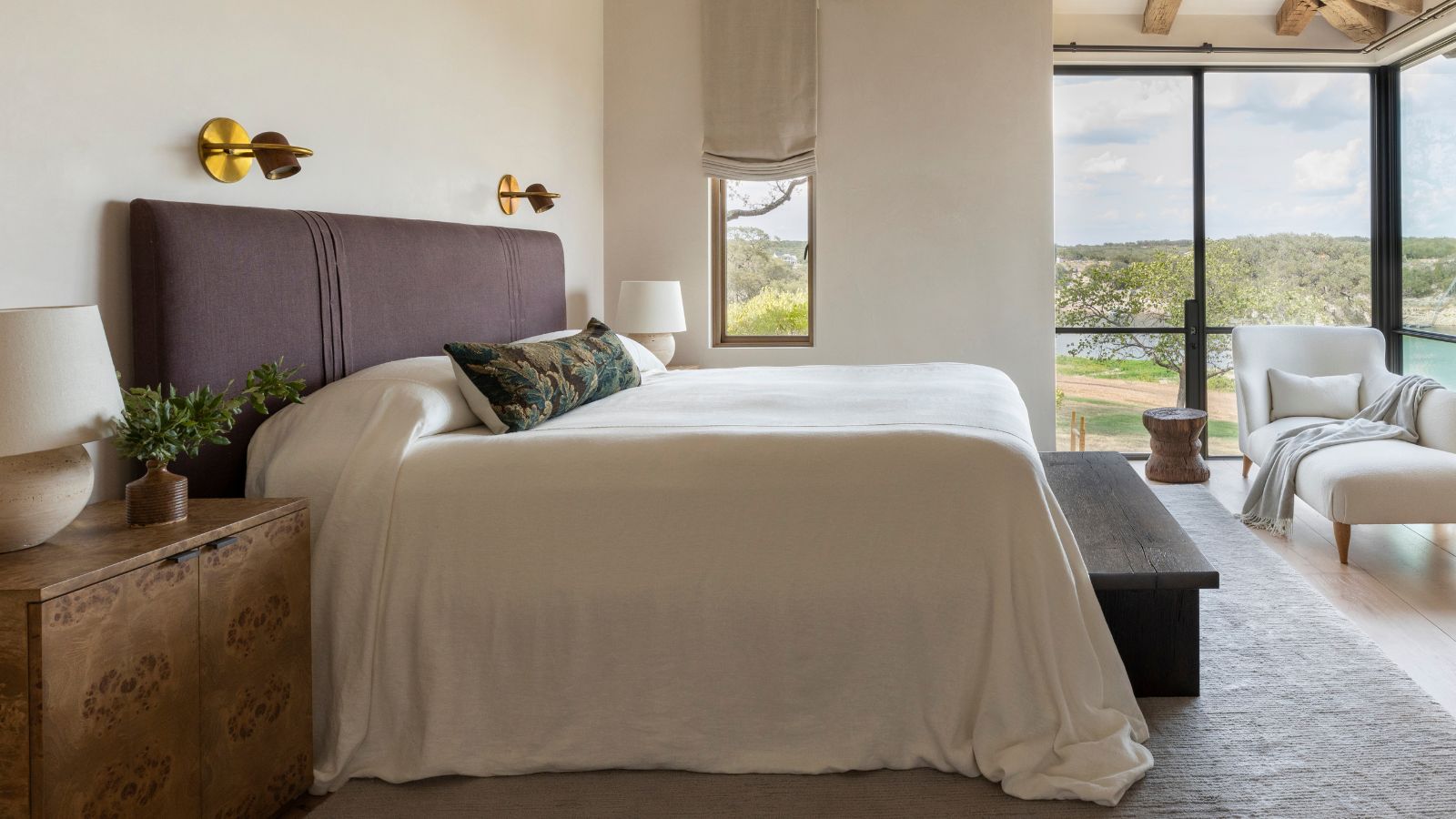 white bedroom with large corner windows, a white chaise lounge, a large bed with a purple upholstered headboard and a burl wood nightstand