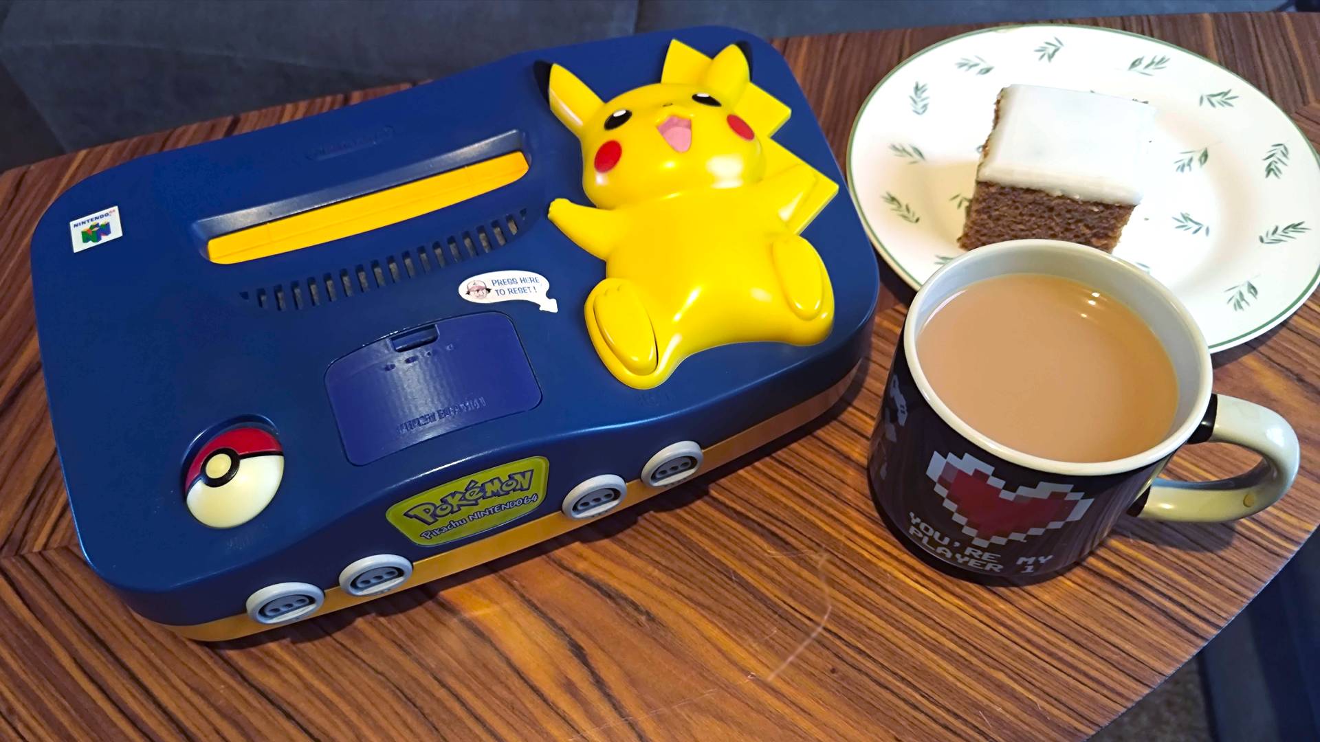 Pikachu N64 next to a cup of tea and iced Ginger cake slice on plate.