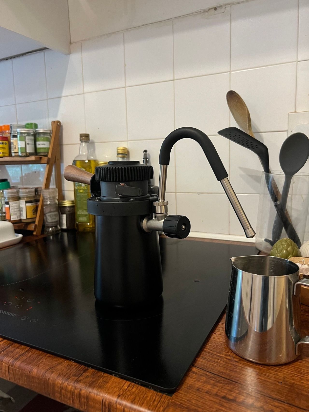 Image of a black stovetop milk steamer on a black hob with a stainless steel milk jug under the steaming spout.