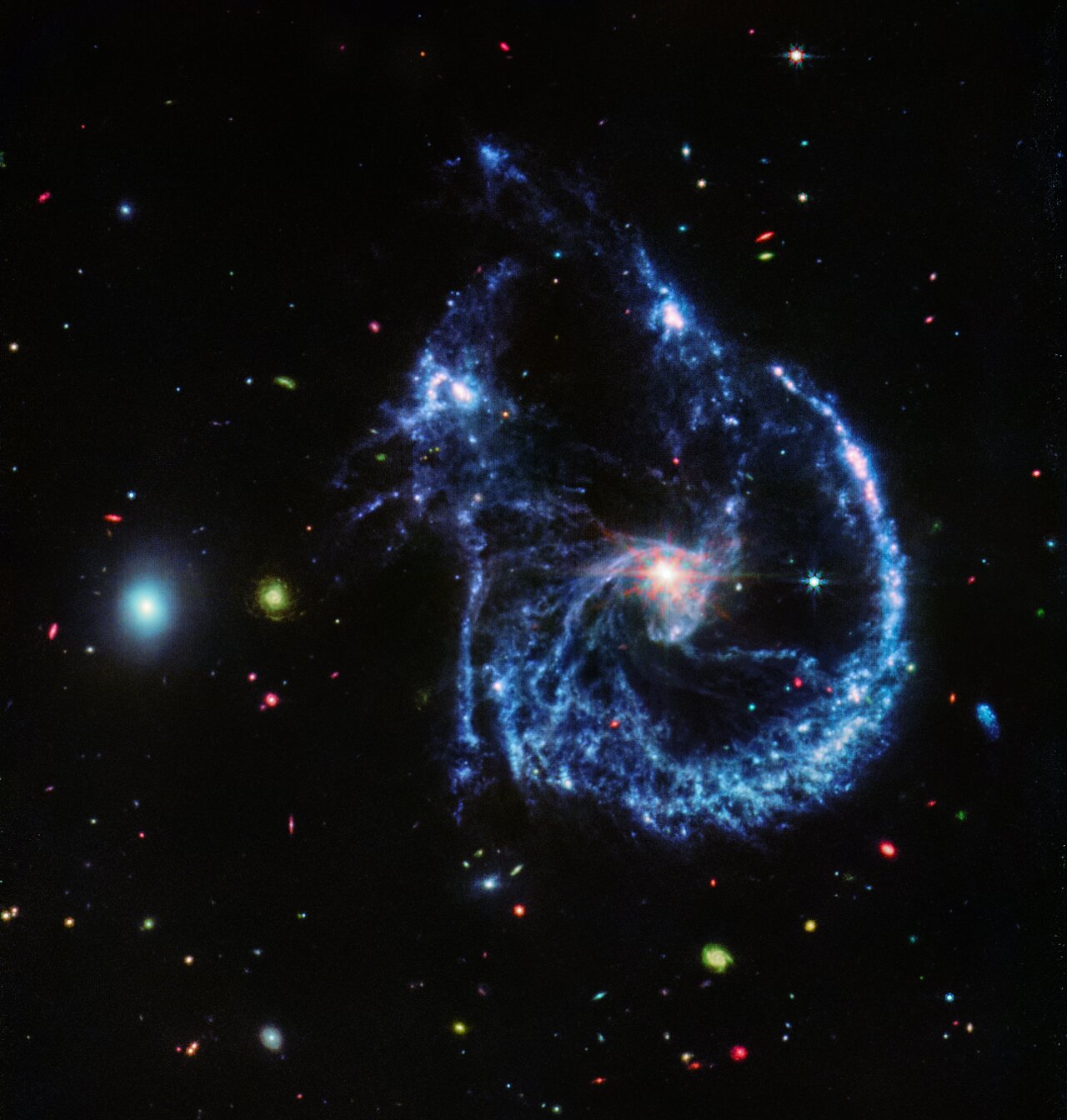 A pair of interacting galaxies. The larger of the two galaxies is slightly right of centre, and is composed of a bright, white centre and a ring of blue, gaseous filaments. The centre of this galaxy shows Webb’s eight-pronged diffraction pattern. There are three filaments of gas and dust moving from the ring toward the centre. At the top left of the ring is a noticeable gap, bordered by two large, blue pockets of dust and gas. The smaller galaxy is made of hazy, light blue gas and dust. Many red, green, blue, and yellow galaxies are spread throughout, with some being hazier in composition and others having more defined spiral patterns